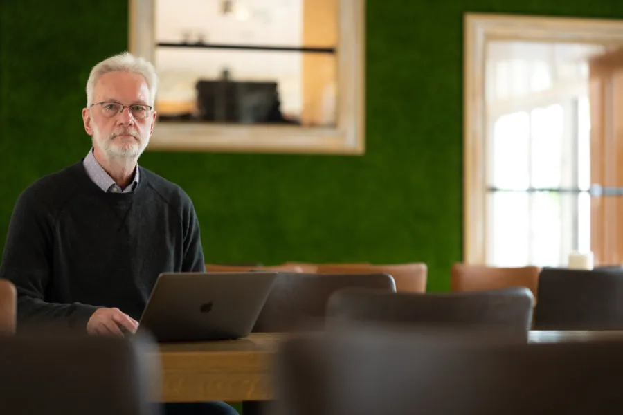 Jürgen Hochhäuser lächelnd mit Laptop an einem Holztisch, im Hintergrund ein Bücherregal
